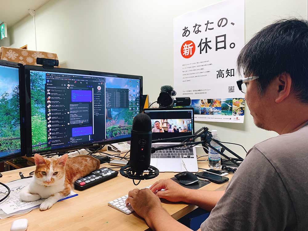 写真：PCに向かう吉冨。机の上にはネコ